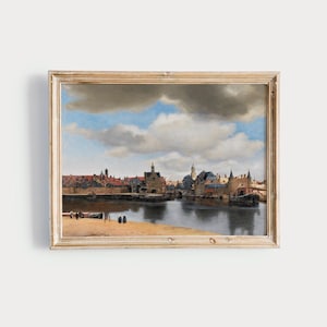 May include: A framed landscape painting depicting a European town with a river, boats, and buildings under a cloudy sky. The artwork is in a gold-colored wooden frame.