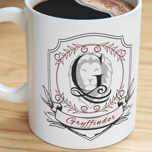 May include: White ceramic mug with a black handle, filled with dark coffee. The mug features a Gryffindor crest design with a lion, the letter "G", and the word "Gryffindor" on a banner. The mug is on a wooden surface.