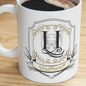 May include: White ceramic mug with a black handle. The mug has a black and gold design with a crest featuring the letter "H" and the words "Hufflepuff".