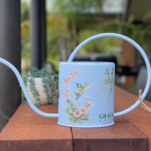 May include: A light blue metal watering can with a curved spout and handle. The can features a hummingbird and floral design with the text "GRACE & AUGUST New Zealand". The watering can is sitting on a wooden surface.