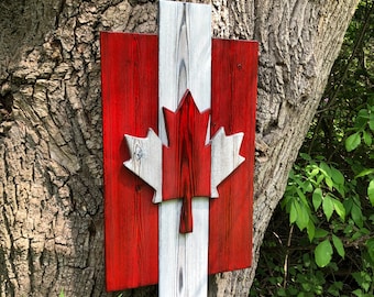 May include: A rectangular wooden wall art piece featuring the Canadian flag design. It consists of two vertical red side panels and a central white panel, with a prominent three-dimensional red maple leaf cutout mounted in the center.