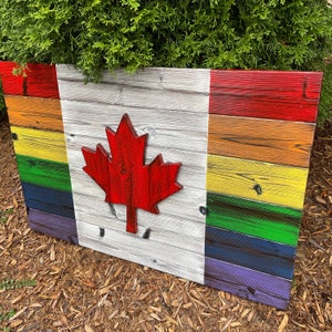 May include: A wooden sign painted with the colours of the rainbow flag and a red maple leaf in the centre. The sign is made of reclaimed wood and has a rustic look.