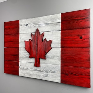 May include: A wooden Canadian flag with a red maple leaf in the centre. The flag is made of reclaimed wood and has a distressed white and red paint finish.