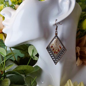 May include: A diamond-shaped earring with a bronze frame and intricate filigree design. The earring features a fan of gold-colored threads and small red beads. The earring is displayed on a white mannequin head, surrounded by greenery and flowers.