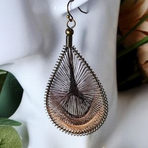 May include: A teardrop-shaped earring with a bronze-colored metal frame and a woven design of brown and tan threads. The earring is suspended from a bronze hook and is displayed on a white mannequin head.
