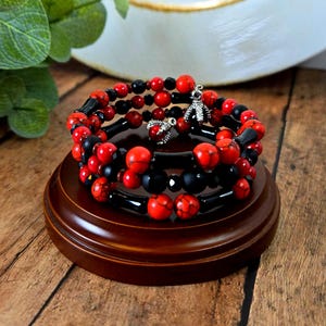 May include: Stacked bracelets featuring red and black beads with silver-toned charms. The bracelets are displayed on a dark wooden stand. The beads have a marbled appearance, and the charms have a detailed design. The bracelets are a stylish accessory.