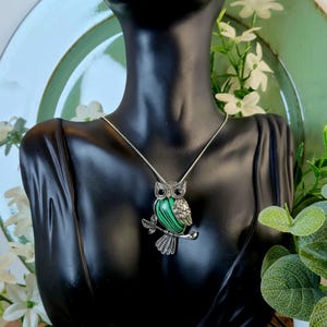 May include: Silver-toned owl pendant necklace. The owl pendant features a green and black striped stone body, detailed silver wings, and sits on a branch. The necklace is displayed on a black mannequin.