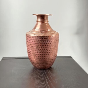 May include: A copper vase with a narrow neck and a wide, rounded body. The surface features a hammered texture with small, repeating indentations across the exterior, complemented by a decorative band of etched patterns around the shoulder area.