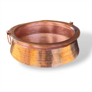 May include: A hammered copper cooking pot with a wide rim and two small handles. The pot has a rounded body and a textured surface. The copper has a warm, reddish-brown color. The pot is designed for cooking and serving.