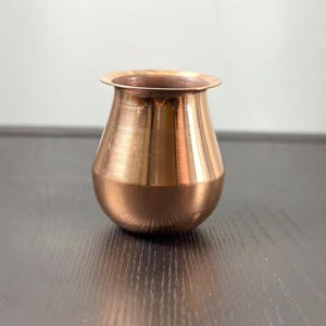 May include: A polished copper vessel with a rounded, bulbous body and a wide, flared rim. The metallic surface features a warm, reddish-brown tone with subtle horizontal striations and a reflective finish that catches the light.