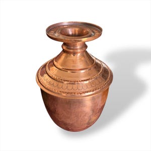 Puede incluir: Un jarrón de color cobre, de estilo antiguo, con una parte superior ancha y plana y un cuerpo bulboso. El jarrón tiene una banda decorativa alrededor del centro y una base acampanada. El metal tiene un tono cálido, marrón rojizo.