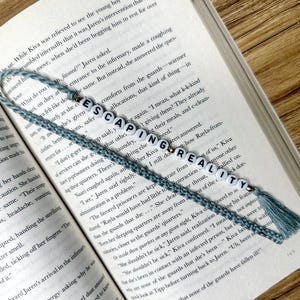 Puede incluir: Un marcapáginas azul y blanco con las palabras "ESCAPING REALITY" en él, descansando en las páginas de un libro. El marcapáginas tiene una borla en el extremo.