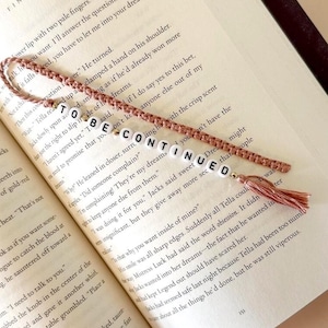May include: A handmade bookmark with the text "TO BE CONTINUED" spelled out in white letter beads. The bookmark has a pink braided cord and a matching tassel, resting on the open pages of a book.