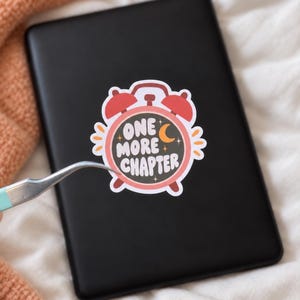 May include: A die-cut sticker shaped like a pink alarm clock with a black face and the white text ONE MORE CHAPTER. The sticker features a small yellow crescent moon and stars, with red bells on top and red legs at the bottom, applied to a black tablet.