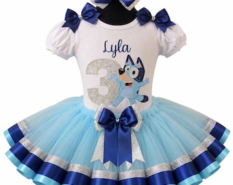 Könnte beinhalten: Ein weißes Kurzarmshirt mit dem Namen Lyla, einer großen silbernen Glitzer-3 und einer blauen Zeichentrick-Hundefigur. Das Shirt hat dunkelblaue Satin-Schleifen an den Schultern und einen hellblauen Tüll-Tutu-Rock mit silbernen und blauen Bändern.
