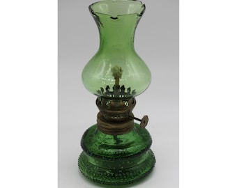May include: A green glass oil lamp featuring a textured hobnail base and a smooth, flared chimney top. The lamp includes a central brass burner mechanism with an adjustable dial and a visible cotton wick centered within the glass globe structure.