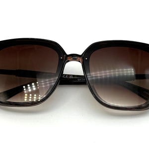 May include: Square-framed sunglasses with a dark brown gradient lens. The frames are a dark brown color with a subtle speckled pattern. The sunglasses are set against a white background.