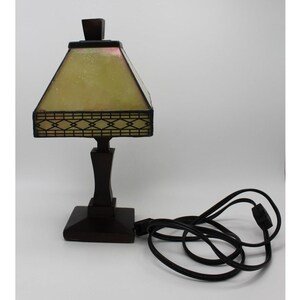 May include: A dark brown table lamp with a square base and a geometric design on the shade. The shade is a light yellow color. The lamp has a black power cord.