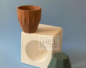 Puede incluir: Un molde para hacer macetas de hormigón. El molde es de forma cuadrada con una abertura redonda en el centro. El molde está hecho de madera y está pintado de blanco. El molde tiene 8,5 cm de alto. La imagen también muestra dos macetas terminadas, una marrón y otra verde.