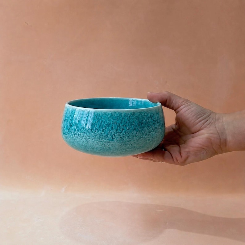 May include: A turquoise ceramic bowl with a textured surface. The bowl is round with a wide opening and a slightly curved rim. The bowl is held by a hand against a peach-colored background.