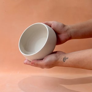 May include: A white ceramic bowl with a rounded shape is held in two hands. The bowl has a smooth, matte finish and a simple, modern design. The background is a solid peach color.