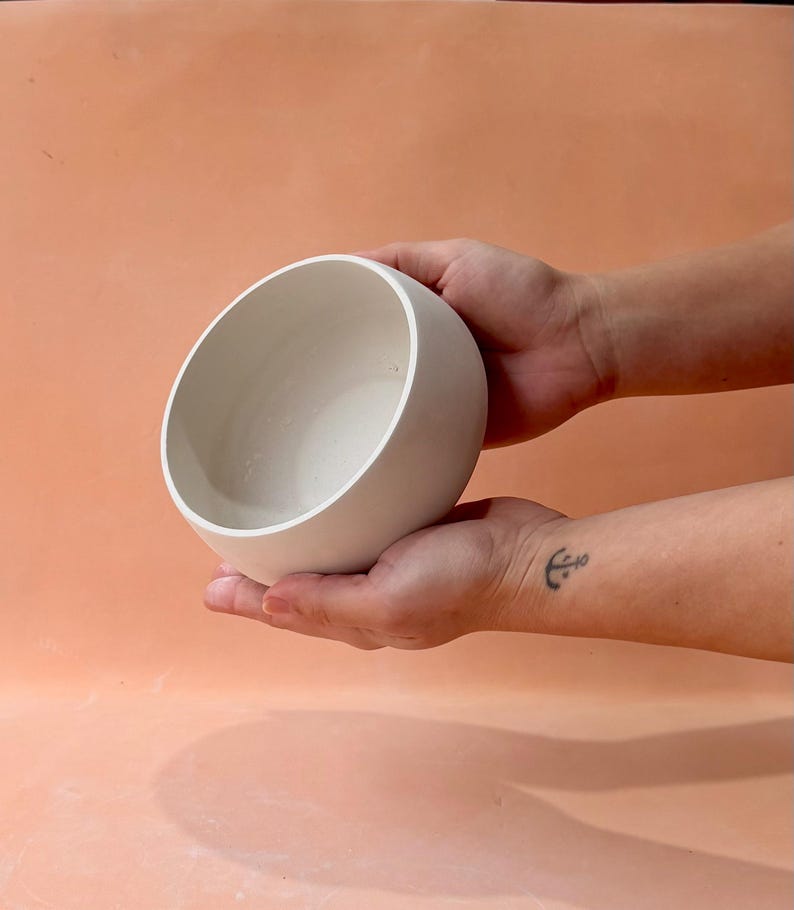 May include: A white ceramic bowl with a rounded shape is held in two hands. The bowl has a smooth, matte finish and a simple, modern design. The background is a solid peach color.