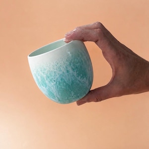 Könnte beinhalten: Eine Hand hält eine Keramiktasse. Die Tasse hat ein einzigartiges Design, das von Weiß oben zu einem türkisfarbenen und weißen Marmormuster übergeht, das an Meereswellen erinnert. Die Tasse hat eine abgerundete Form und eine glatte Oberfläche.