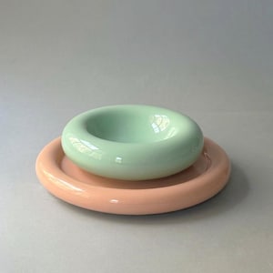 May include: A smooth, glossy ceramic set consisting of a pale mint green doughnut-shaped bowl resting on a larger, flat, circular peach-coloured plate. Both pieces feature rounded edges and a uniform, reflective finish with no visible patterns.