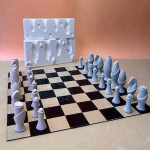 Puede incluir: Un juego de ajedrez con piezas abstractas de cerámica gris sobre un tablero de ajedrez negro y beige. Las piezas están dispuestas en posición inicial. Dos moldes de cerámica blanca se encuentran en el fondo.
