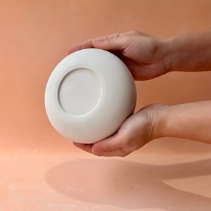 May include: A white, round ceramic bowl with a recessed center, held by two hands. The bowl has a smooth, matte finish and a simple, minimalist design. The bowl is likely used for serving or display.