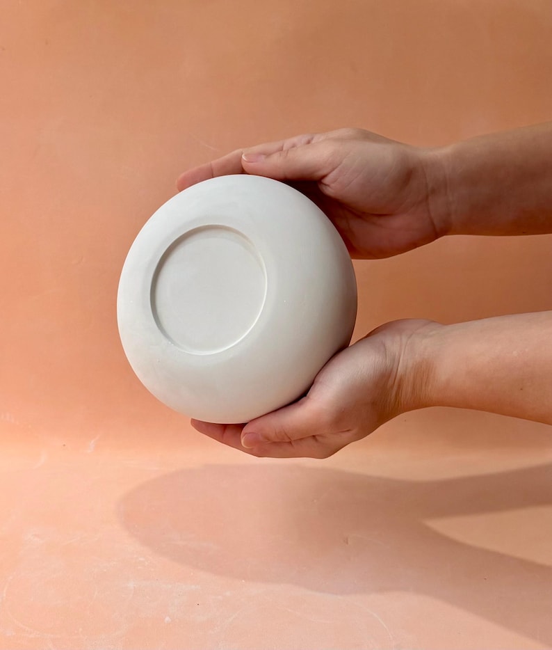May include: A white, round ceramic bowl with a recessed center, held by two hands. The bowl has a smooth, matte finish and a simple, minimalist design. The bowl is likely used for serving or display.