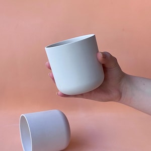 Puede incluir: Dos macetas cilíndricas de cerámica gris claro. Una se sostiene en una mano, la otra descansa sobre una superficie color melocotón. Las macetas tienen un acabado liso y mate y una base redondeada. El interior es un tono de gris más oscuro.