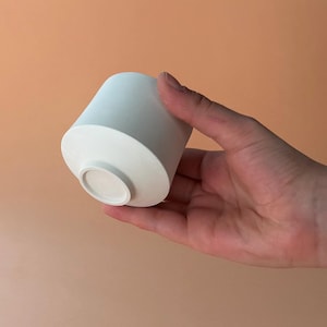 Op de afbeelding: Een kleine, witte keramische beker met een cilindrische body en een licht uitlopende bovenkant. De beker wordt in een hand gehouden tegen een perzikkleurige achtergrond. Het ontwerp van de beker is minimalistisch en modern.