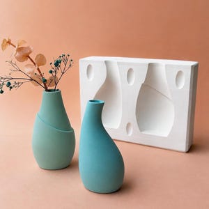 Può includere: Due vasi in ceramica nei toni del blu-verde e del turchese, con uno stampo bianco sullo sfondo. Il vaso a sinistra ha un design diagonale. I vasi sono su una superficie rosa, con fiori secchi nel vaso a sinistra.