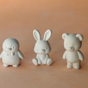 May include: Three unpainted ceramic figurines: a penguin with a bow tie, a seated bunny, and a standing teddy bear. The figures are white and have smooth surfaces, ideal for painting or decorating.