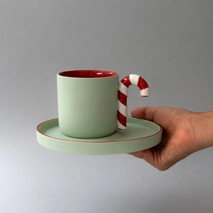 Puede incluir: Un juego de taza y platillo de cerámica en color verde claro. La taza cuenta con un asa en forma de bastón de caramelo con rayas rojas y blancas. El interior de la taza y el borde del platillo tienen un acabado en rojo intenso, contrastando con el exterior verde claro mate de ambas piezas.