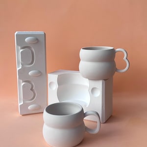 May include: Two white ceramic mugs with rounded bodies and unique handle designs. One mug sits atop a white square form, while another rests on a white rectangular mold. The background is a solid peach color.