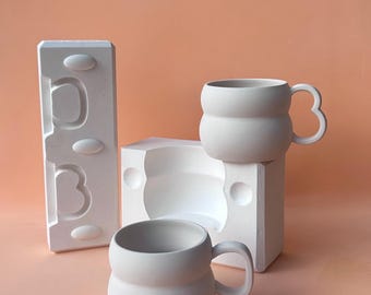 Puede incluir: Dos tazas de cerámica blanca con cuerpos redondeados y diseños de asa únicos. Una taza está sobre una forma cuadrada blanca, otra sobre un molde rectangular blanco. El fondo es de color melocotón sólido.
