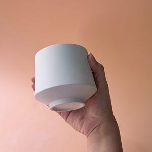 Può includere: Un vaso di ceramica bianco, cilindrico, con un design geometrico. Il vaso ha una base ampia e piatta e una parte superiore leggermente conica. La finitura liscia e opaca contrasta con lo sfondo color pesca.
