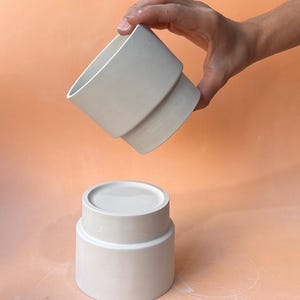 Puede incluir: Un juego de dos recipientes de cerámica blanquecina. La pieza superior se sostiene sobre la base, revelando un diseño escalonado. La base es cilíndrica con una parte superior plana. El fondo es de color melocotón sólido.