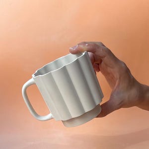 May include: A white ceramic mug with a unique, fluted design. The mug has a handle and a wide base. The mug is held against a peach-colored background.