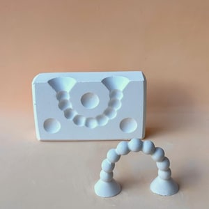 May include: A white, rectangular mold with a curved beaded design and a separate, matching beaded arch. The mold has two large cone-shaped openings and three circular indentations. The arch is made of small, round beads.