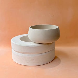 May include: A light beige ceramic bowl sits atop a stack of two white, circular ceramic forms. The bowl has a rounded shape with a wide opening. The background is a solid peach color.
