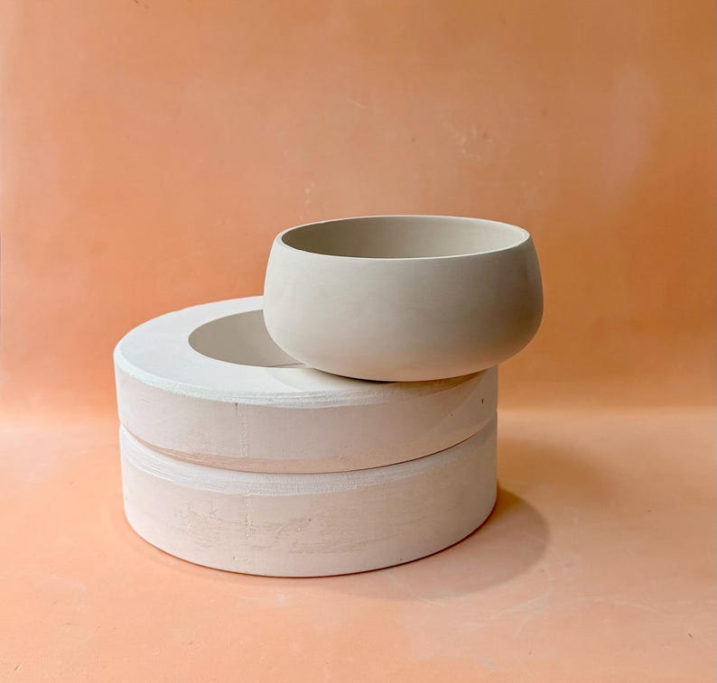 May include: A light beige ceramic bowl sits atop a stack of two white, circular ceramic forms. The bowl has a rounded shape with a wide opening. The background is a solid peach color.