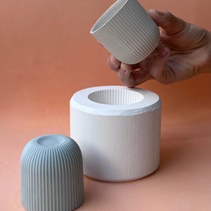 Puede incluir: Tres piezas de cerámica de estilo minimalista. Una pequeña taza acanalada azul claro se encuentra sobre un fondo melocotón. Una taza acanalada blanca más grande se sostiene en una mano sobre un recipiente cilíndrico blanco con una abertura central.