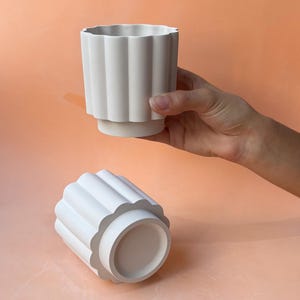 Puede incluir: Dos macetas de cerámica blanca con un diseño acanalado. Una maceta está en posición vertical, sostenida por una mano, mientras que la otra está tumbada de lado. Las macetas tienen un borde festoneado y una base de pedestal. El fondo es de color melocotón sólido.