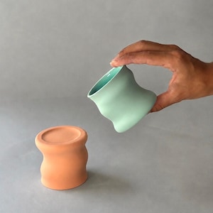 May include: Two ceramic cups with a distinct hourglass-like, wavy silhouette. One cup is a matte terracotta orange color, while the other is a smooth, light mint green color with a matching mint green interior finish.
