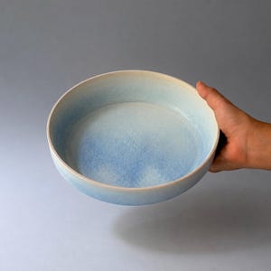 Pode incluir: Uma tigela redonda de cerâmica com um interior liso de cor clara que transita para um acabamento exterior azul degradado e salpicado. A tigela possui uma borda circular limpa e uma base larga e rasa.