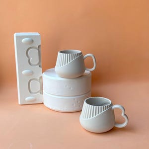 Könnte beinhalten: Zwei cremeweiße Keramikbecher mit einzigartigem Design, einer auf einem weißen Podest. Die Becher haben eine abgerundete Form mit einem diagonal gerippten Abschnitt. Eine weiße Form mit herzförmigem Design ist ebenfalls vorhanden, vor pfirsichfarbenem Hintergrund.