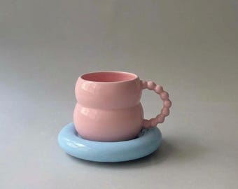 Puede incluir: Una taza de cerámica rosa claro con un cuerpo redondeado y abombado, y un asa compuesta por una serie de pequeñas esferas. La taza descansa sobre un platillo circular azul claro liso con un borde grueso y redondeado, creando un contraste.